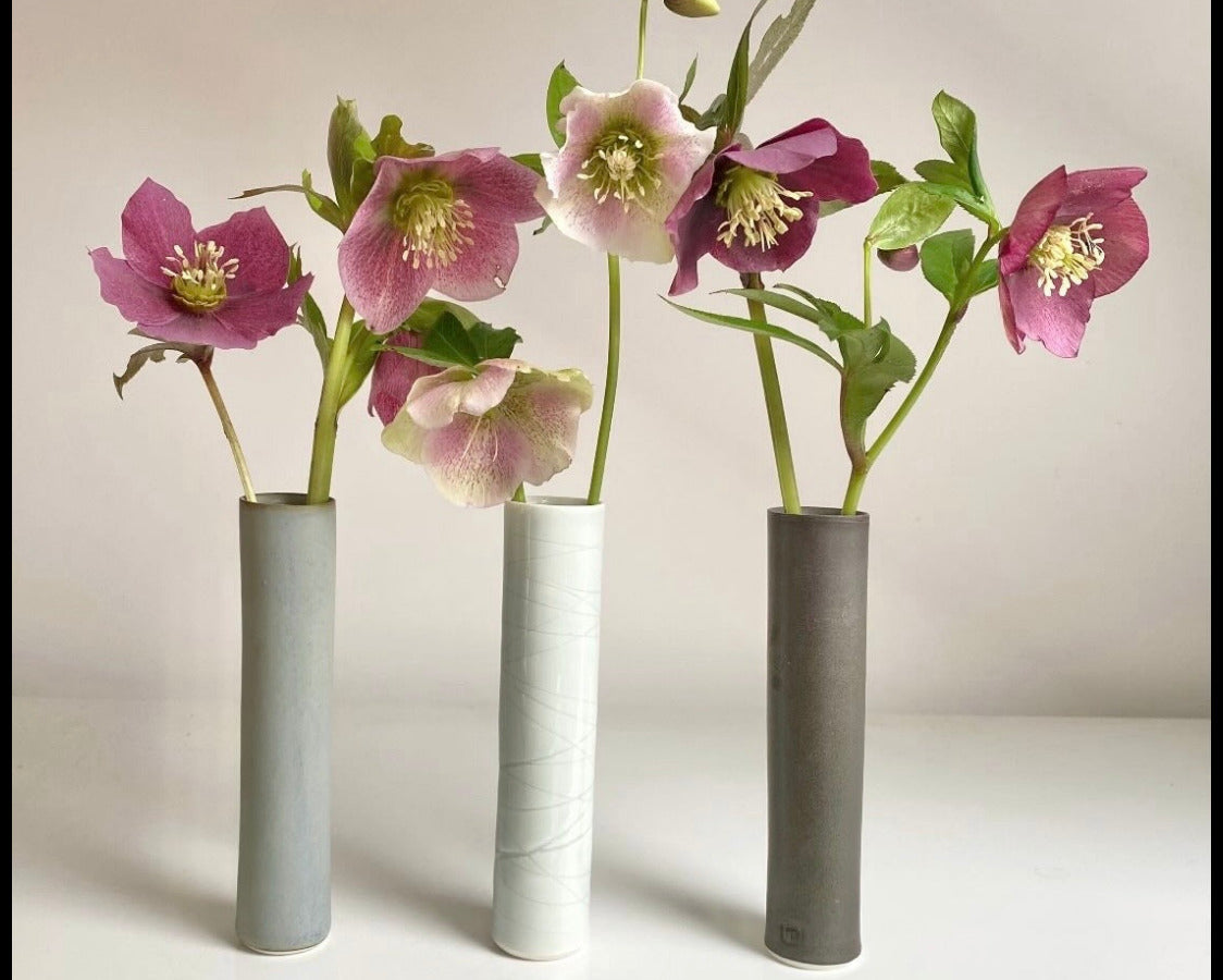 Three minimalist cylindrical ceramic vases in light blue, white, and dark gray, each holding a single pink hollyhock flower with green leaves, arranged on a white surface against a light beige background.